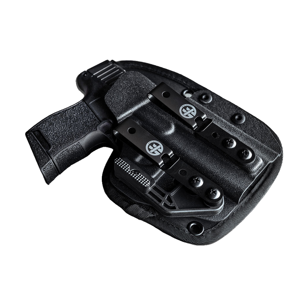 A black polymer holster containing a black handgun, shown from an angled top-down view against a transparent background. The holster features a quick-release clip with a circular crosshair logo and a textured grip on the pistol.